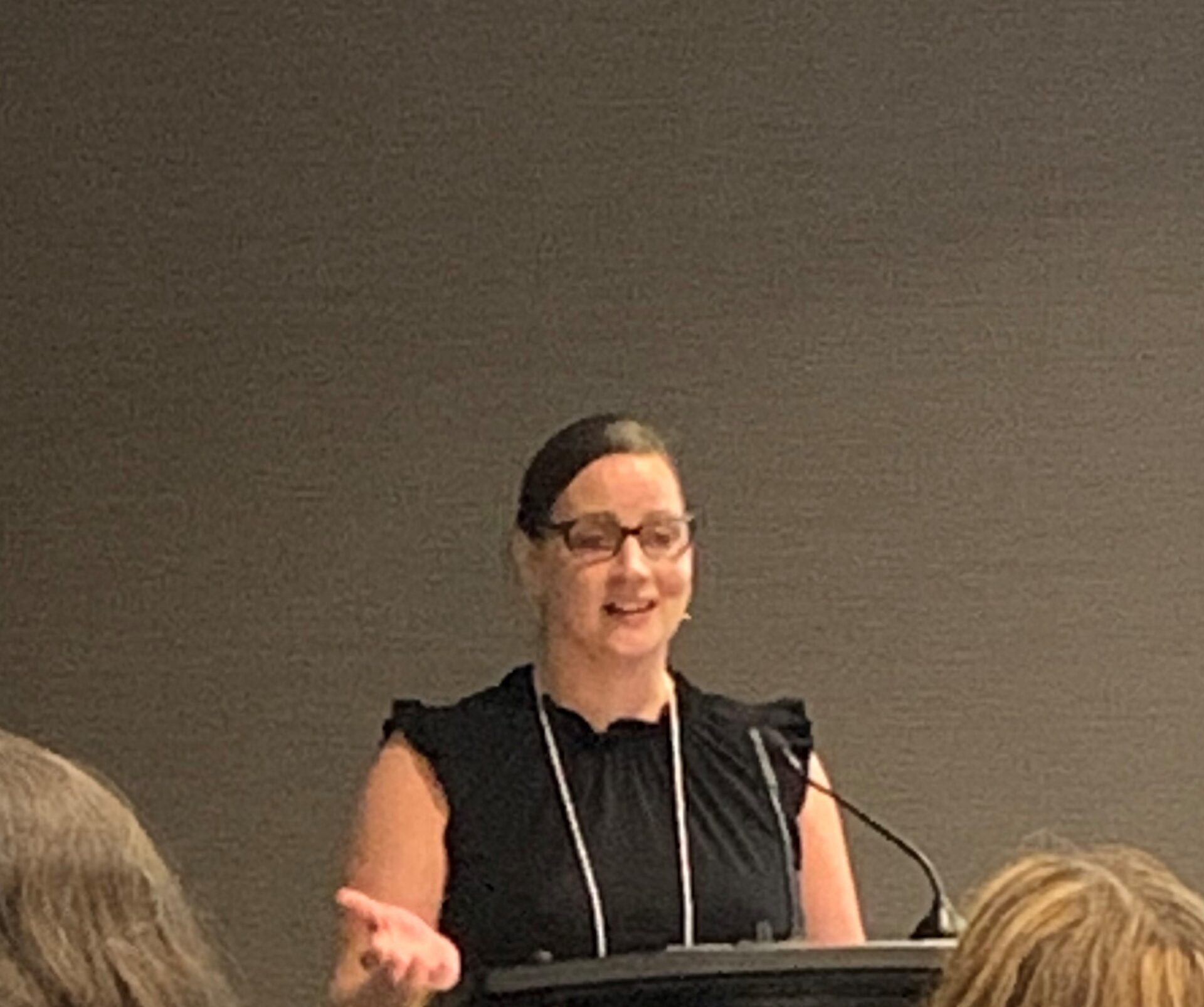 Kailey Bilodeau stands at a podium, dark hair pulled back into a pony tail, dark rimmed glasses, wearing a black top with a lanyard over it.