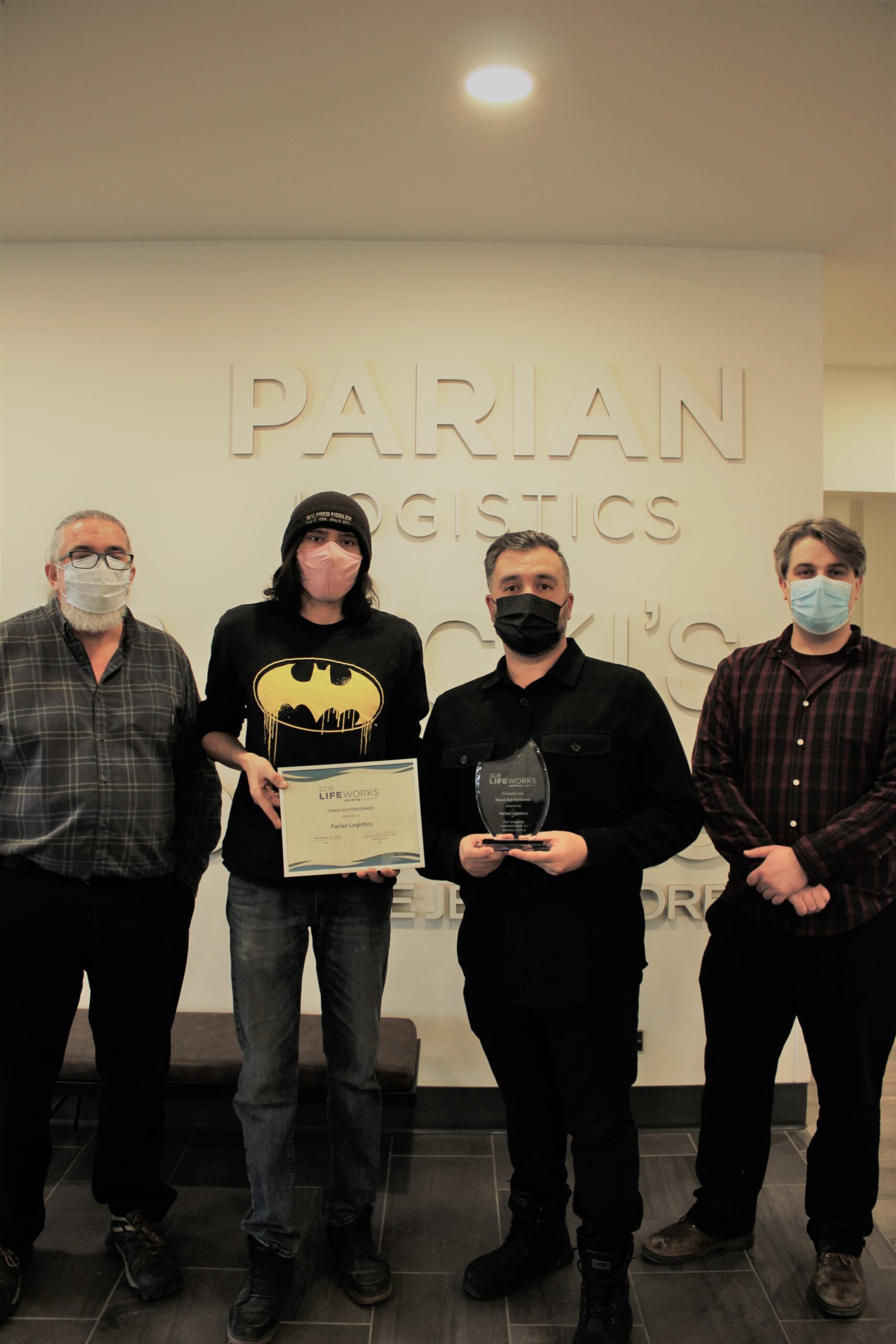 four people stand in a row in front of a sign that says Parian Logistics, they are wearing face masks and holding awards and ceritificates.