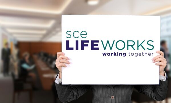 Out of focus office with a person standing up front holding a whiteboard sign and the SCE LifeWorks logo is on the sign