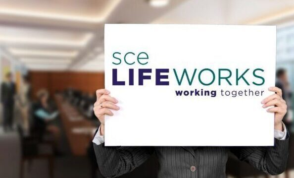 Out of focus office with a person standing up front holding a whiteboard sign and the SCE LifeWorks logo is on the sign