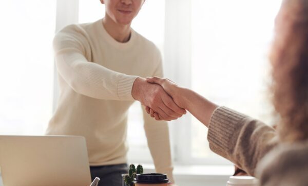 Two people shaking hands over a desk one, standing and one seated.