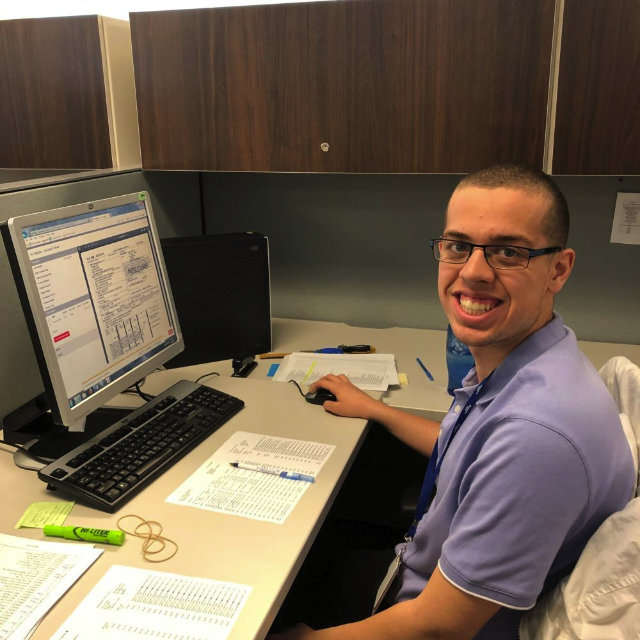 Young smiling man sits in front of a computer. He has dark very short hair and eyeglasses, wearing a violet blue shirt