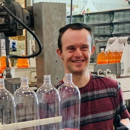 Mike in shorts and striped shirt standing behind bottles on conveyor belt.